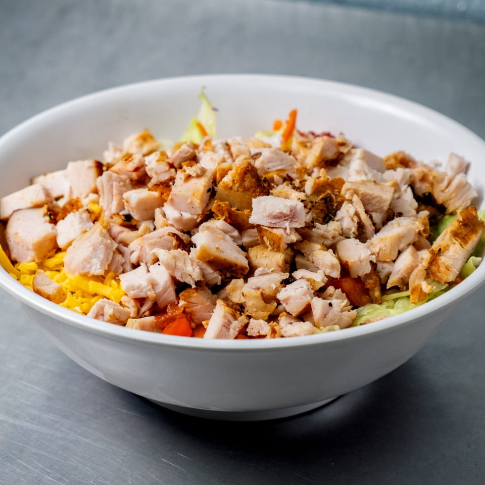 A white bowl filled with a salad topped with generous chunks of grilled chicken, cheese, and vegetables.